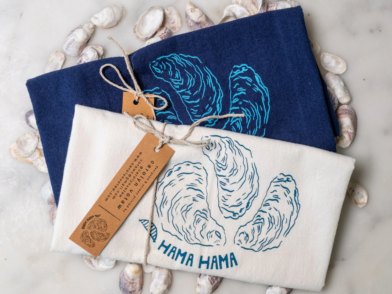 Two tea towels with oyster designs and 'Hama Hama' text on a stone surface with shells.